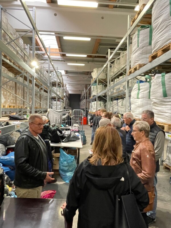Eine Personengruppe steht vor großen Regalen gefüllt mit in großen Säcken verpackten Kleidungsstücken